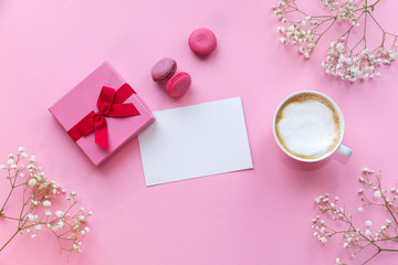 Flatlay pink coral background, the cup of cappuccino coffee and sweets macaroons, spring white flowers