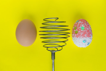 One colorful decorated easter egg and one real egg and whisk on yellow pastel color background. Easter cooking concept, greeting card.