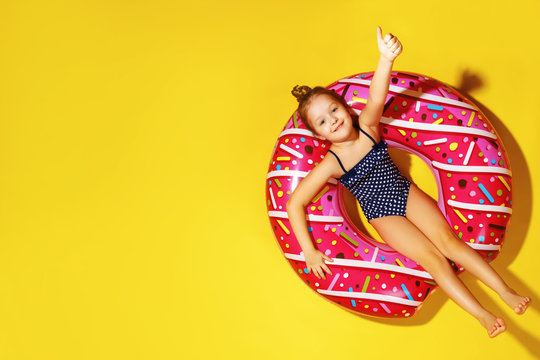 Little girl in a bathing suit, lying on a donut inflatable circle. A child shows thumbs up. Yellow background. Top view. Summer concept.