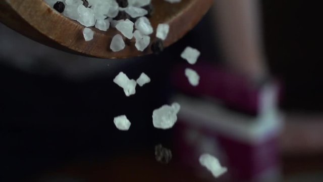 Coarse salt and black pepper are poured from a wooden bowl in slow motion side view