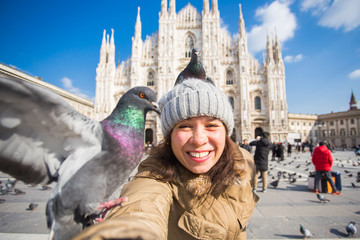 Fototapeta na wymiar Italy, excursion and travel concept - young funny woman taking selfie with pigeons in front of cathedral Duomo in Milan