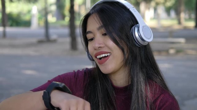 Modern pretty asian woman with headphones talking with her smart watch-outdoor