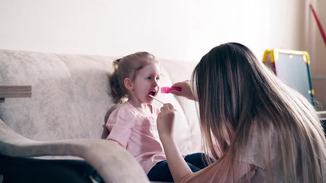 4k: Young attractive mother and sweet daughter play the role of doctor and patient. The daughter opens her mouth, her mother looks into her throat, flashing a flashlight.