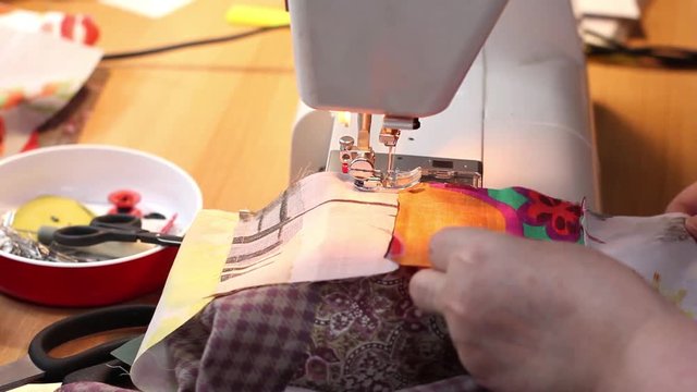 Mature woman is stitching by sewing machine part of patchwork quilt, directing fabric under foot by hands. Close-up.