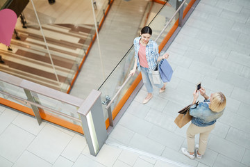 Top view of blond lady using smartphone to photograph friend with shopping bags in modern mall