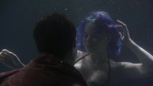 The blue-haired mermaid and the dark-haired guy swim together under the dark water among the bubbles and look at each other. Close-up portrait.
