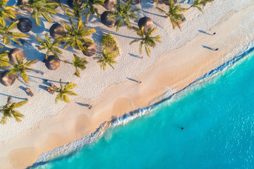 Aerial view of umbrellas, palms on the sandy beach, people, blue sea with waves at sunset. Summer...