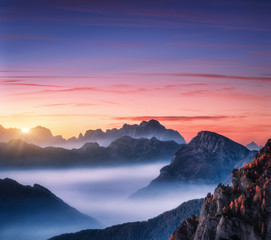 Mountains in fog at beautiful sunset in autumn in Dolomites, Italy. Landscape with alpine mountain valley, low clouds, trees on hills, red sky with clouds at dusk. Aerial view. Passo Giau. Travel