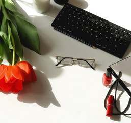 Women's office desk top view.Female workspace with laptop, flowers red tulips, glasses, make up products on white background with sunlight shadows. Copy space. Holiday concept