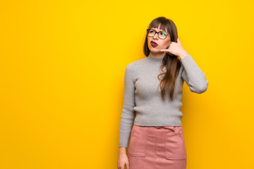 Woman with glasses over yellow wall making phone gesture and doubting