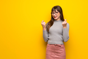 Woman with glasses over yellow wall making money gesture