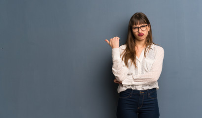 Woman with glasses over blue wall unhappy and pointing to the side