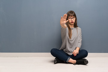 Woman with glasses sitting on the floor making stop gesture denying a situation that thinks wrong