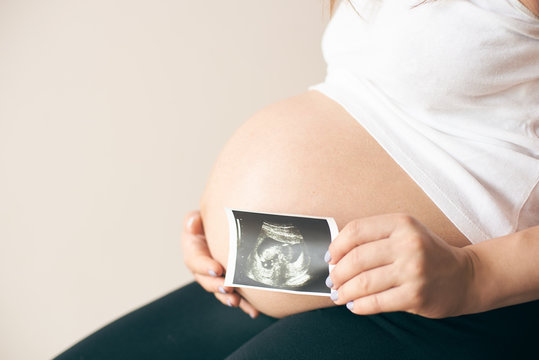 Side view of pregnant woman in white shirt sitting and showing ultrasound image of baby. Future young mother expecting child. Concept of anticipation and parental love.