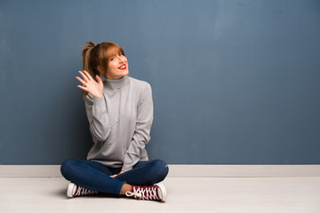 Redhead woman siting on the floor saluting with hand with happy expression