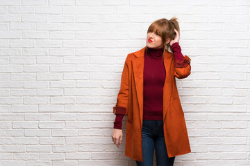 Woman with coat over white brick wall having doubts while scratching head