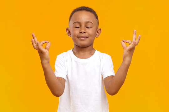 Peaceful calm little boy of African appearance wearing casual t-shirt keeping his eyes closed and smiling, meditating isolated in studio. Conscious dark skinned schoolboy practicing meditation