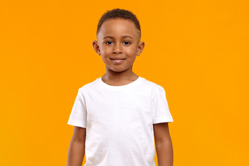 Isolated image of cute adorable dark skinned schoolboy wearing white t-shirt posing in blank yellow...