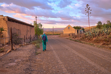Karoo dirt road leading to a bend in morning light. Man in green overall by farm house seen from...