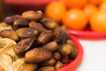 Dried fruits and dates lie on a plate on a light wooden background. In the background are tangerines. copyspace