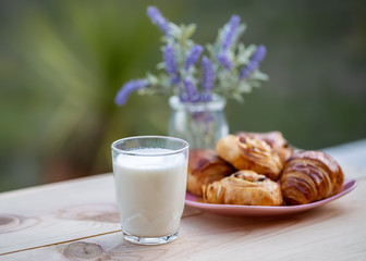A glass of milk on rustic table. Buns with raisins and french croissant. Dinner in an orchard in summertime. Bouquet of lavender. Dessert. Soft focus. Summer twilight. Blurry background. Copy space.