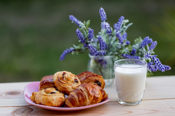 Buns with raisins and french croissant. A glass of milk on wooden table. Dinner in an orchard in summertime. Bouquet of lavender. Dessert. Soft focus. Summer twilight. Blurry background. Copy space