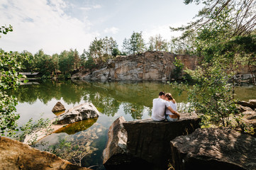 Man and woman sit back and hug on stone near lake on background of big rocks. Canyon. Place for text and design. Reflected. Landscape of an old flooded industrial granite quarry filled with water.