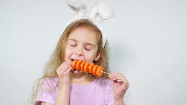 The girl cheerfully nibbles carrot pieces on a stick. Child with bunny ears eating carrot.