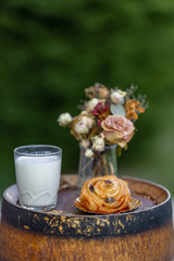 Bun with raisins and a glass of milk on old wine barrel. Dessert. Bouquet of withered flowers. Dinner in an orchard in summertime. Summer twilight. Soft focus. Blurry background. Closeup
