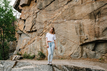 Slim girl stands and poses at cliff. Portrait of a woman in jeans and a white T-shirt, against the background of the mountain on nature. Full length. Looking at the camera. Close up