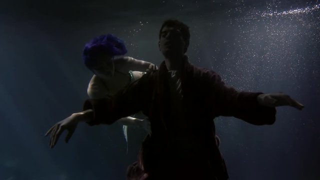 A young man with his eyes closed, slowly falling under the water and circling around him, swims a beautiful mermaid girl with blue hair