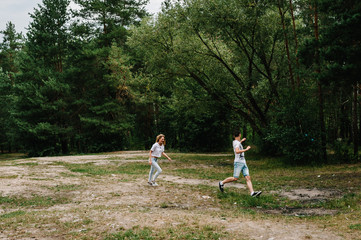Fototapeta premium Young couple running away and having fun on the background on the nature. A woman catches her husband against the background of trees and fir trees.