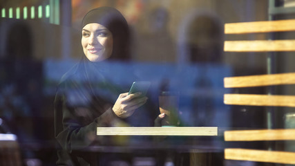 Beautiful Muslim lady holding coffee and smartphone in hands, sitting in cafe