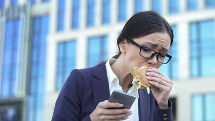 Young female manager eating unhealthy burger in hurry, checking news on phone