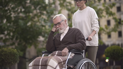 Young woman on walk with sad pensioner in wheelchair, caregiver assistance, help