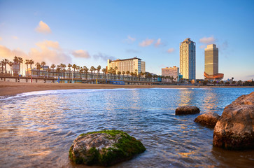 Obraz premium Barcelona, Spain. Panoramic view at Barceloneta beach from sea. View with stones. Sunset landscape with blue sky. Sandy coastline with palms and waves of surf. Popular touristic vacation destination.
