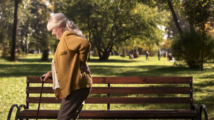 Fototapeta premium Senior lady slowly walking with stick along park suffering from back pain