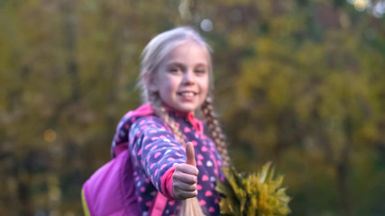 Beautiful schoolgirl with autumn leaves looking at camera and showing thumbs up