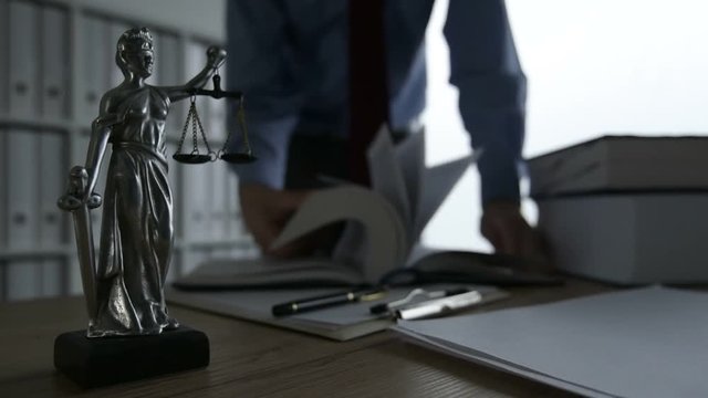 Lawyer flipping pages of the law book in office, selective focus on statue of Lady Justice