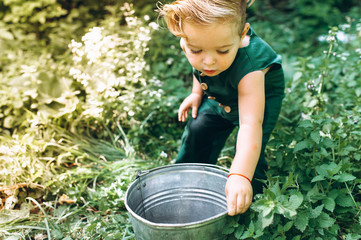 A boy with white hair in a green combination plays with a bucket on the grass