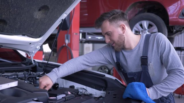 car service, attractive technician guy checks oil level during automobile repair with open hood at auto workshop, smiles and gives positive gesture