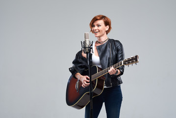 Fototapeta premium Portrait of a young woman musician with an acoustic guitar. Sing a song on studio microphone on a gray background. She wears black leather jacket and plays rock and roll loudly. Space for text