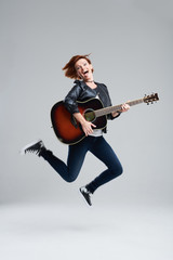 Young woman musician with an acoustic guitar in hand on a gray background. She laughs and jumps high. plays rock and roll loudly. Full-length portrait. 