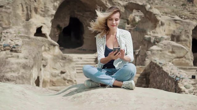 Tourist woman sits on stone checks mail on phone holding in hands. freelancer works. Background ancient city carved into white rocks Uplistsikhe Georgia. happy smling face adult girl, positive emotion