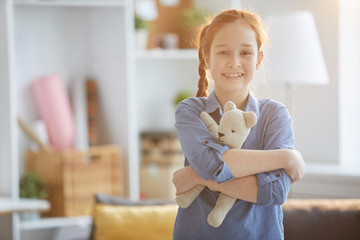 Waist up portrait of happy red haired girl hugging plush toy and looking at camera lit by warm...