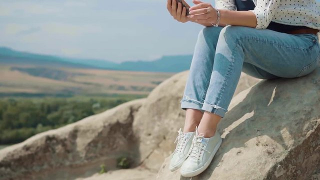Cute girl blogger sitting on a stone against the sky, valley and mountains. In touch with his subscribers talking on the phone and broadcasts the details of his fun. Casual wear jeans, shirt, sneakers