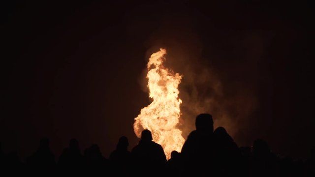 Massive bonfire with bright orange flames leaping into the sky lights up people gathered around it. Nighttime celebration with a raging fire on the beach. People in silhouette stand near a huge fire.