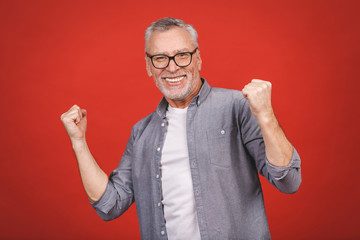 Happy mature senior man in casual using smartphone and making winner gesture isolated over red...