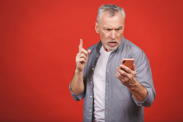 Portrait of angry senior man talking on mobile phone isolated on red wall background. Negative...