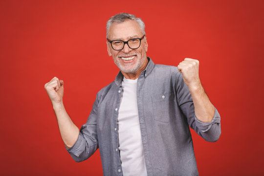 Happy Mature Senior Man In Casual Using Smartphone And Making Winner Gesture Isolated Over Red Background.